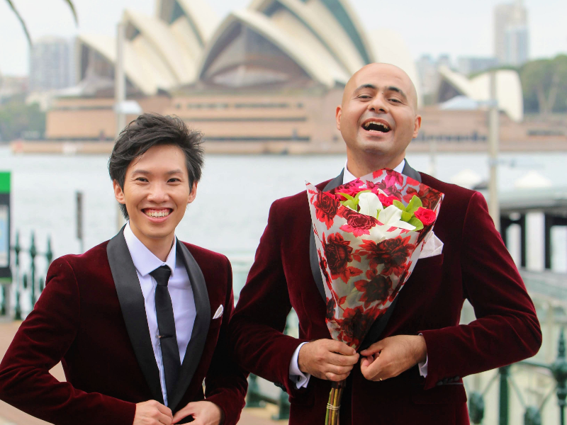 two men dressed in red velvet suits holding a bouquet in front of the Sydney opera house after being granted Australian citizenship